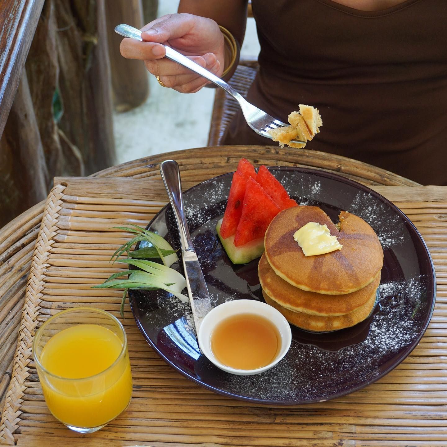A stack of pancakes with butter, a side of watermelon, a small bowl of syrup, and a glass of orange juice on a tray.