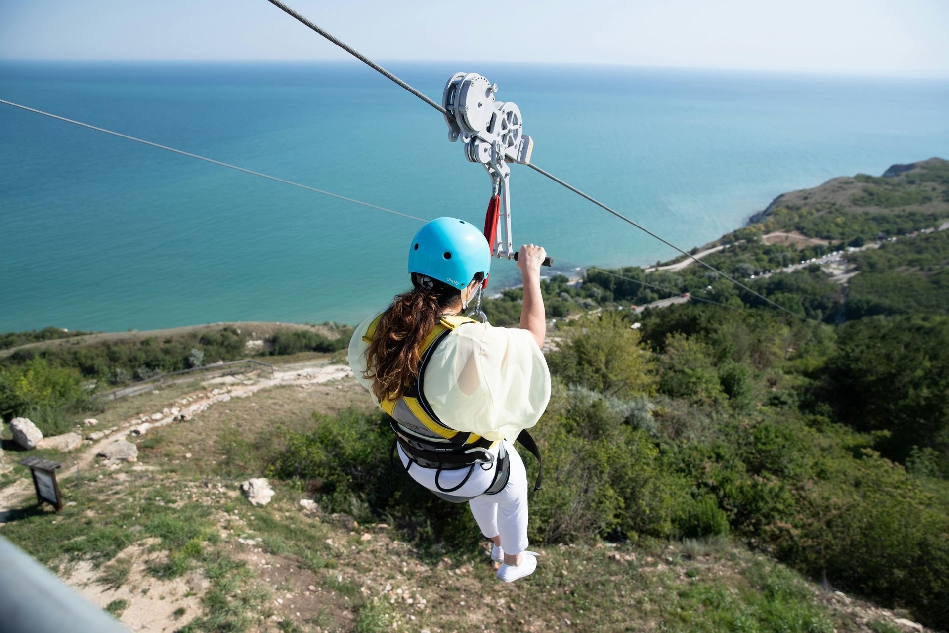 A person in a blue helmet and white shirt rides a zipline over a green hillside with a view of the sea.
