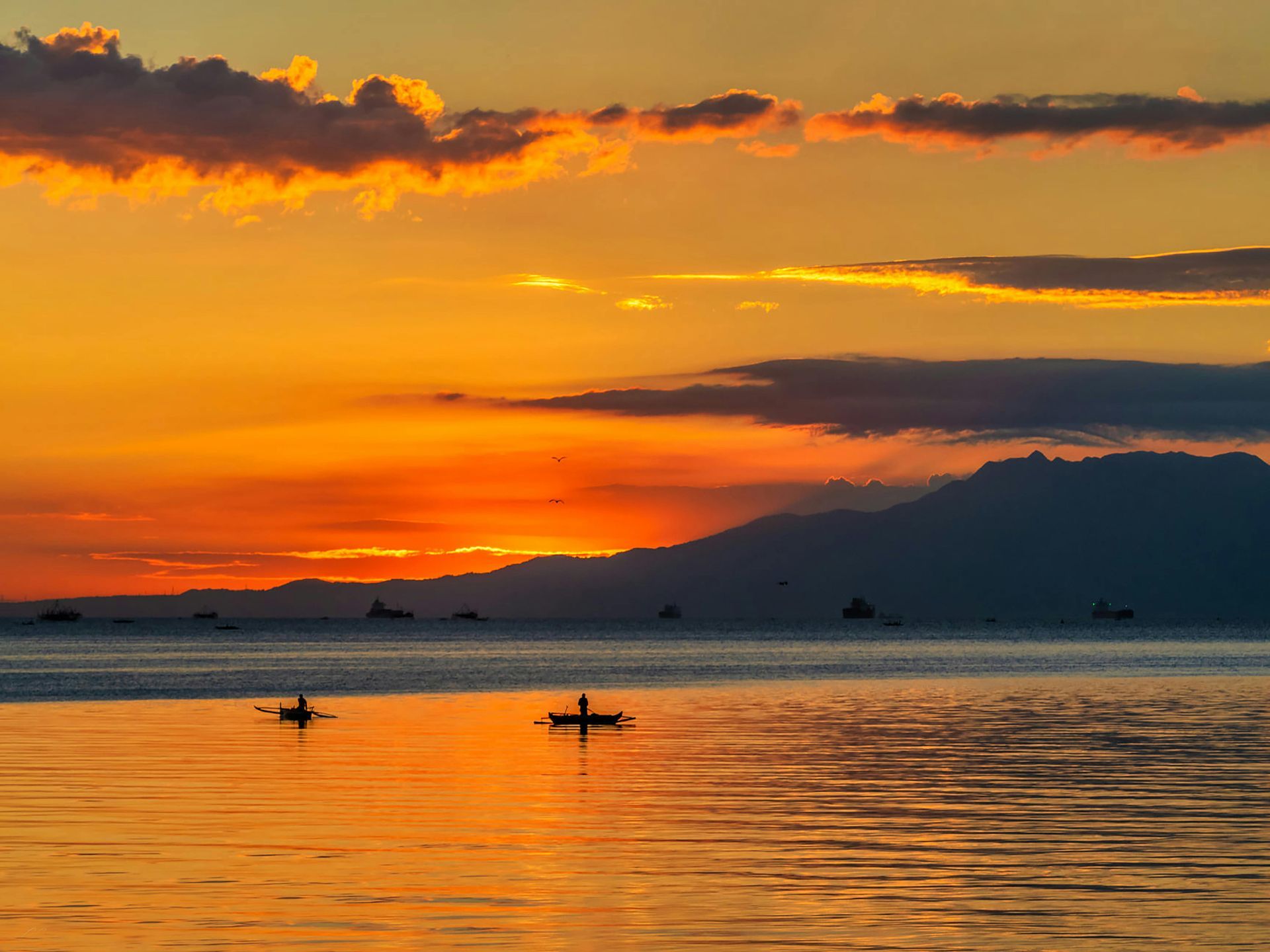 Two small boats float on calm, reflective water against a vibrant orange sunset, with a dark mountain silhouette behind.