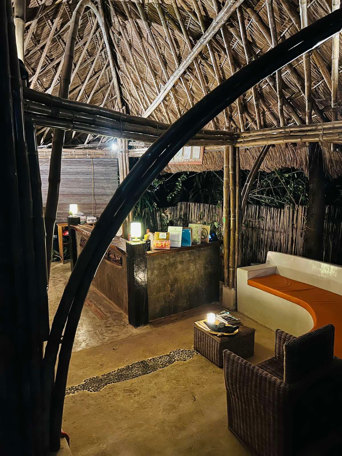 An open-air lounge with a thatched roof, featuring a wicker chair, a bench with an orange seat, and a small service bar.