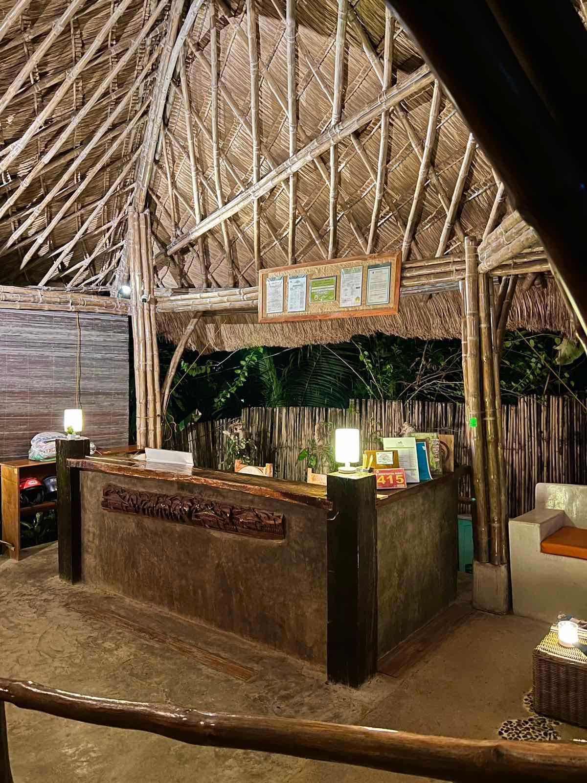 A rustic, thatched-roof reception desk area with warm lighting, wood accents, and a sign in a dimly lit, natural setting.