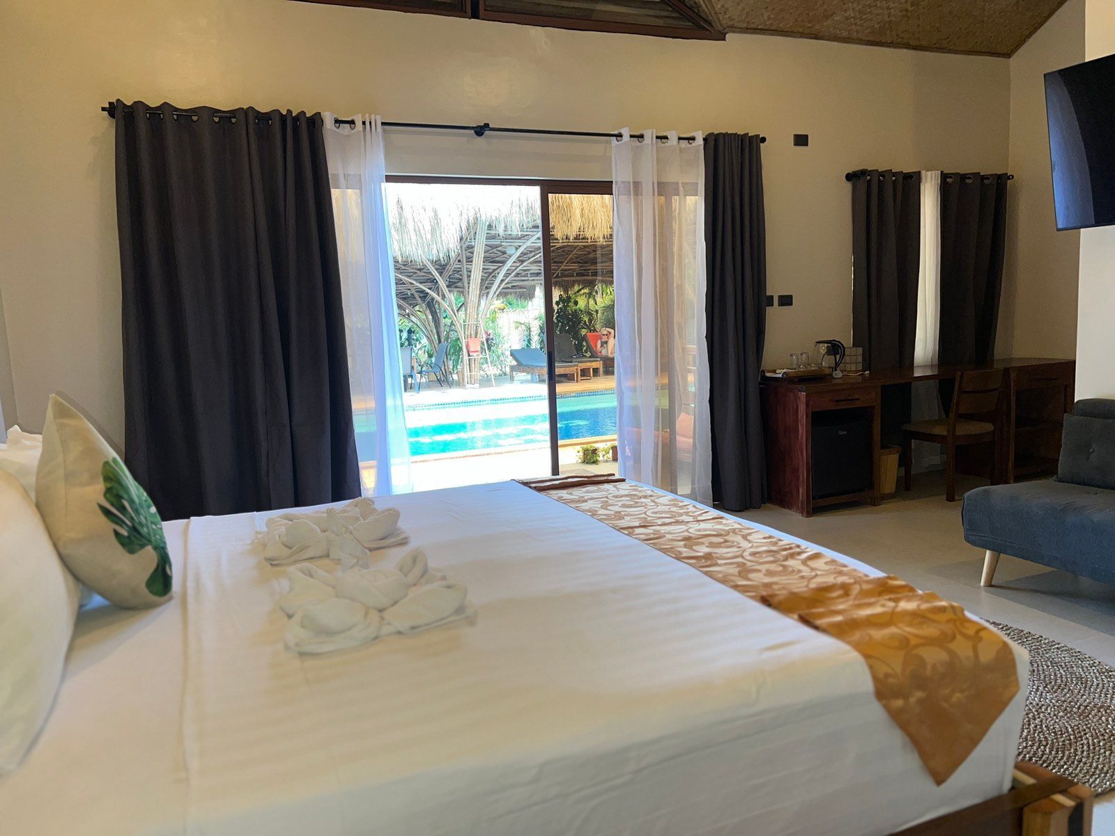 A bedroom with a bed featuring white linens and a gold runner, facing a window view of a pool with patio seating.