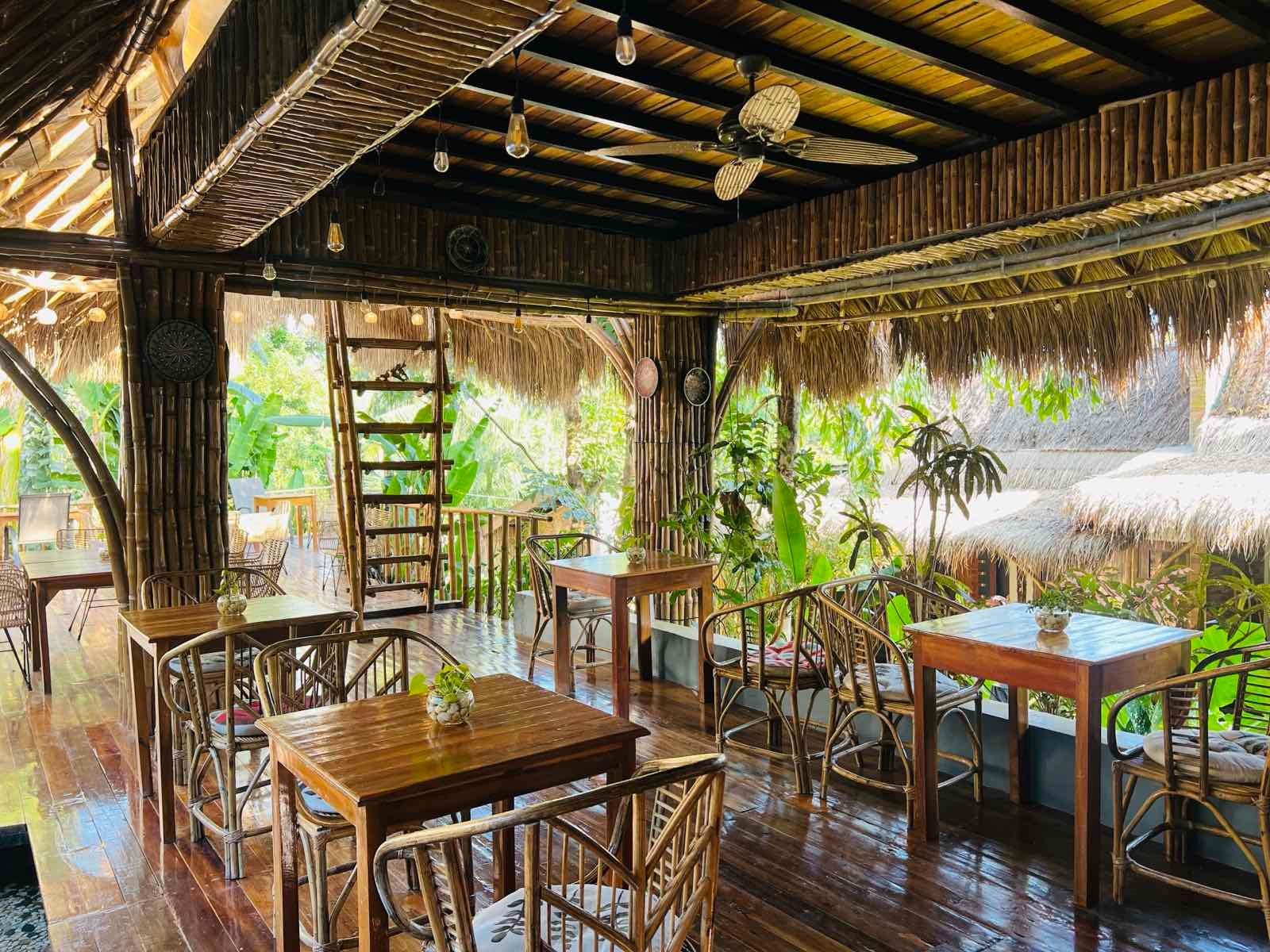 A wooden, open-air restaurant terrace with tables, chairs, and a thatched roof overlooking lush tropical greenery.
