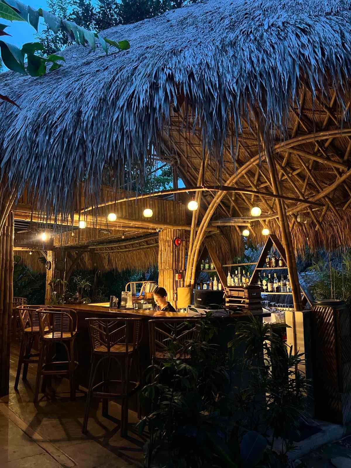 A tiki-style open-air bar at dusk, featuring a thatched roof, warm string lights, and wooden bar stools.