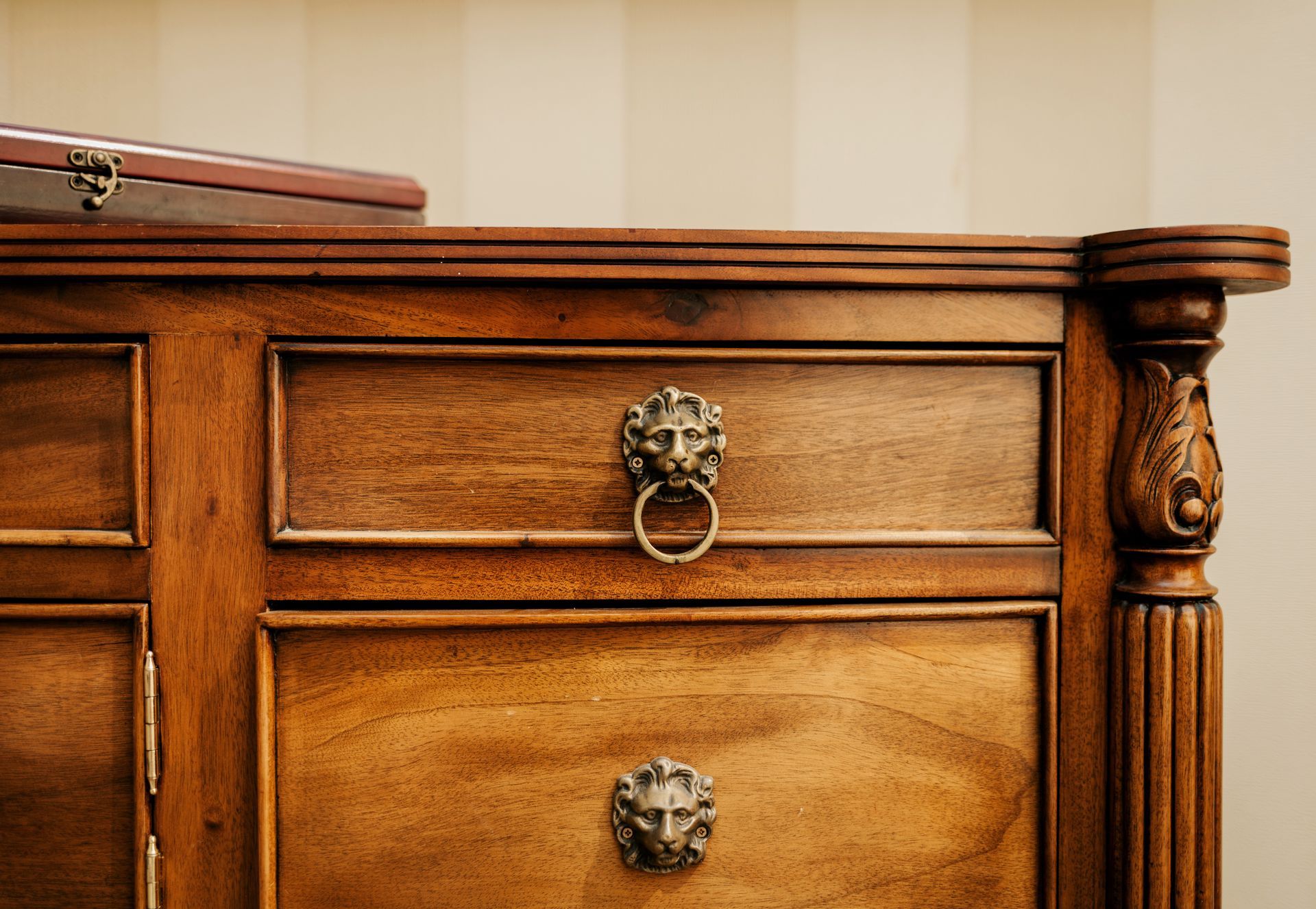 Close-up of a wooden dresser featuring two drawers with bronze, lion-head ring pulls and an ornate carved column.