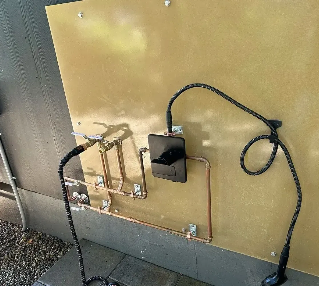 An outdoor shower system with copper pipes and a handheld showerhead mounted on a tan panel attached to a grey wall.