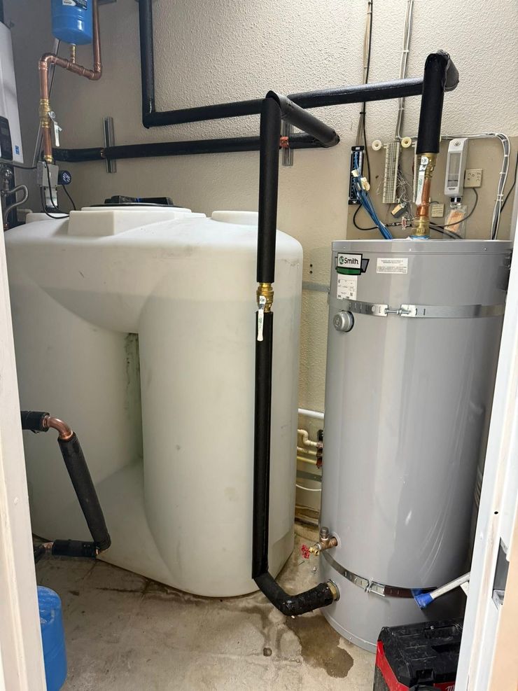A white plastic water storage tank stands next to a gray cylindrical water heater, both connected by insulated pipes.