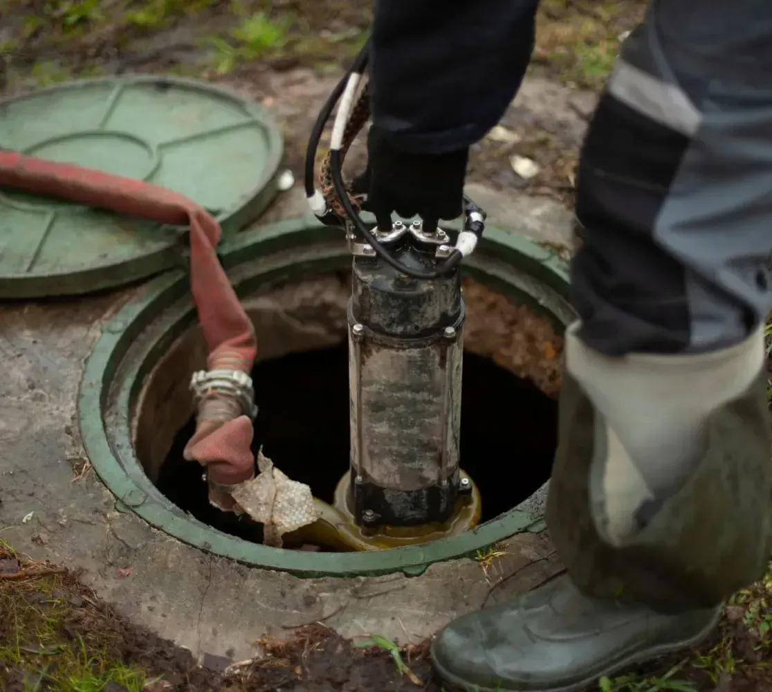 A worker uses a submersible pump and hose to empty a dark, open septic tank in an outdoor setting.