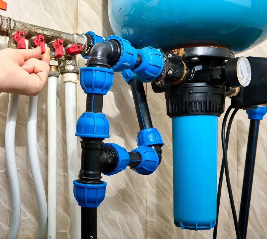 A hand adjusts a red valve on a plumbing manifold connected to a blue water filtration system against a tiled wall.