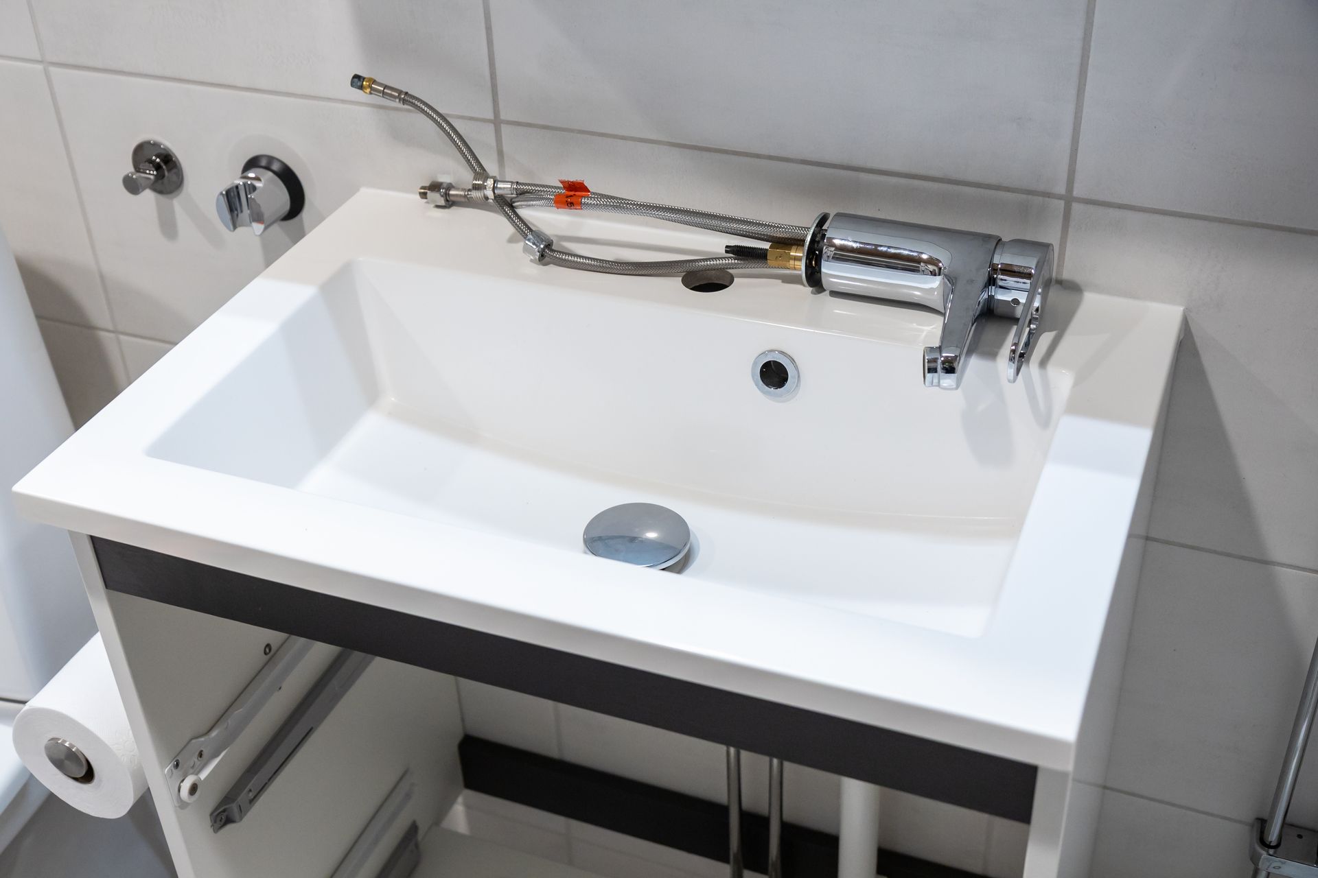 A modern bathroom sink with a disassembled metal faucet lying on its edge, set against a tiled wall.
