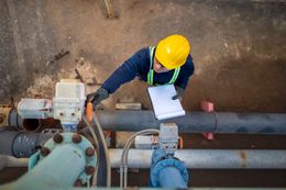 An inspector in a yellow hard hat and high-vis vest checks industrial pipes while holding a notepad.