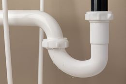 A white P-trap pipe under a sink with small water droplets dripping from the bottom curve.