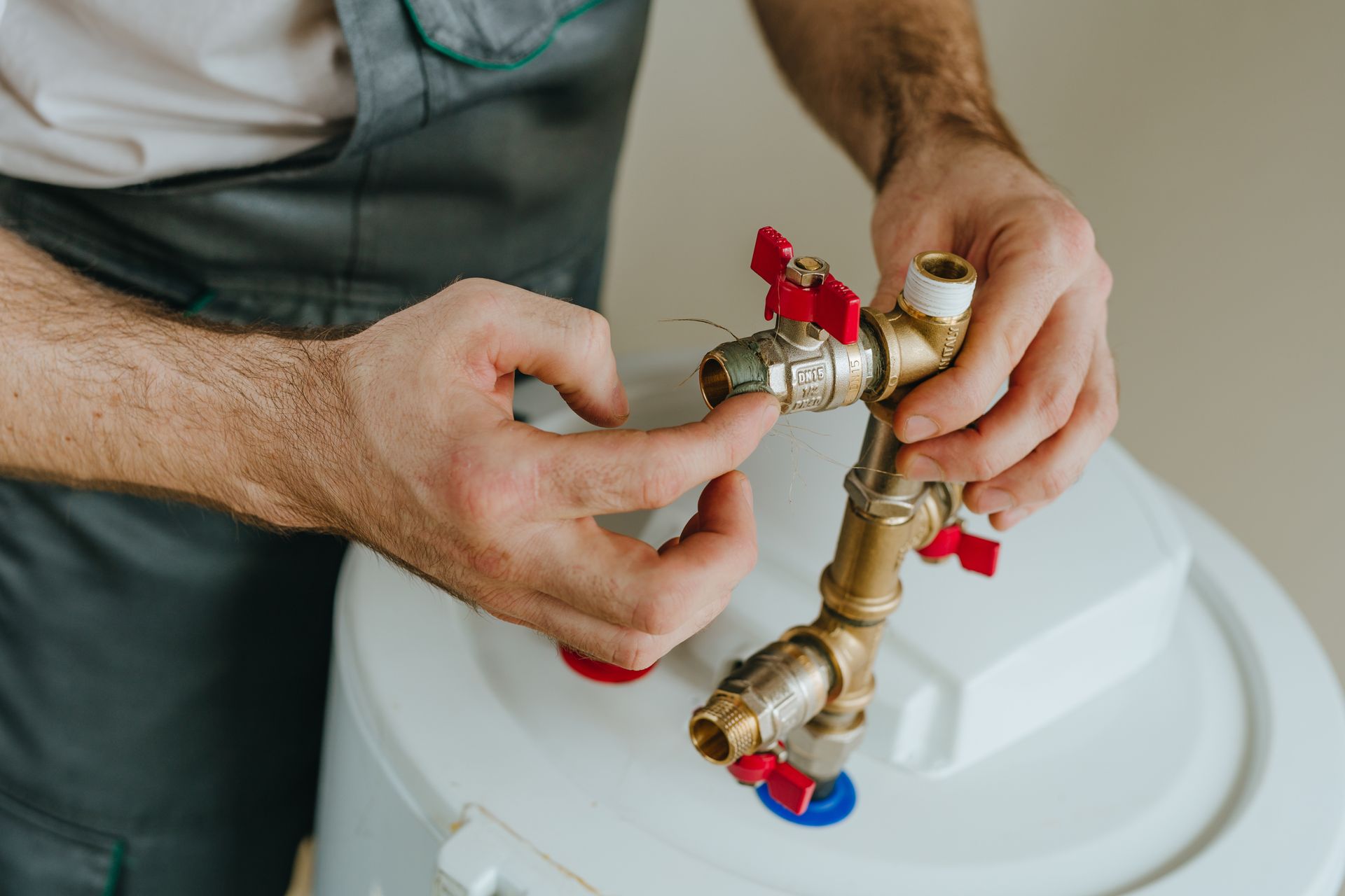 A plumber in grey workwear assembles brass pipe fittings with red valves on top of a white water heater.