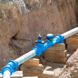 A blue industrial water pipe fitting installed in a dirt trench, supported by wooden blocks.
