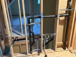 Black plumbing pipes are installed within an unfinished wooden wall frame during construction.