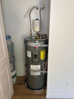 A gray water heater with a small expansion tank sits inside a closet, secured to the wall with metal strapping.
