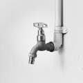 A metal water faucet with a T-shaped handle attached to a white pipe against a plain, light-colored wall.