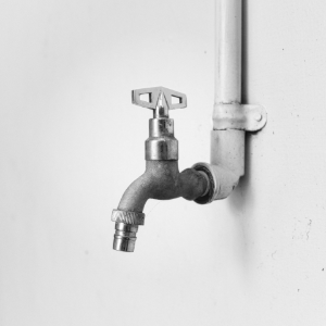 A metal water faucet with a T-shaped handle attached to a white pipe against a plain, light-colored wall.