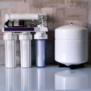 A white reverse osmosis water filtration system with three filter canisters and a storage tank sits on a reflective surface.
