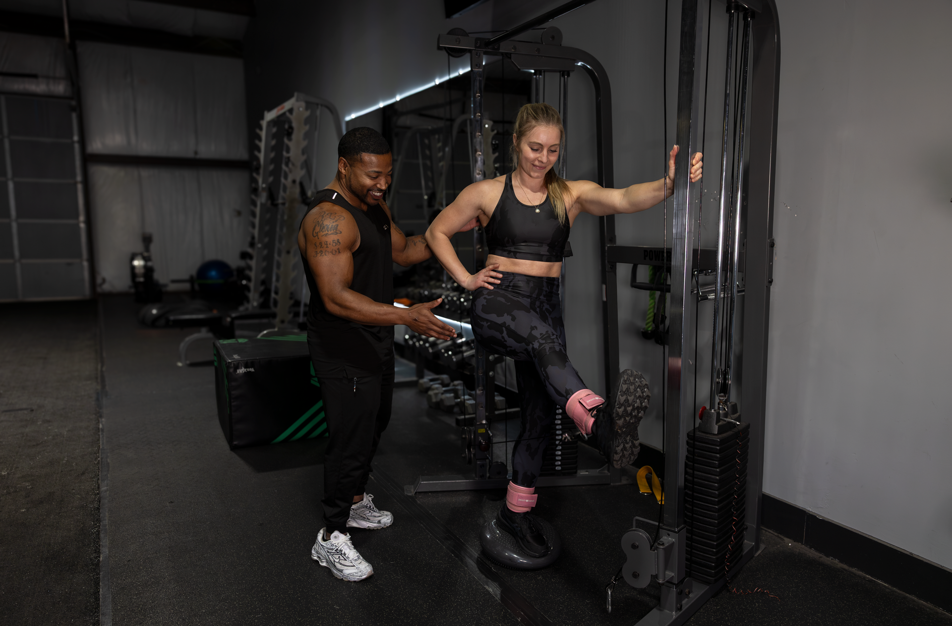 360 image fitness trainer Trey Johnson assists female client with leg strength exercises to help build muscle wearing black