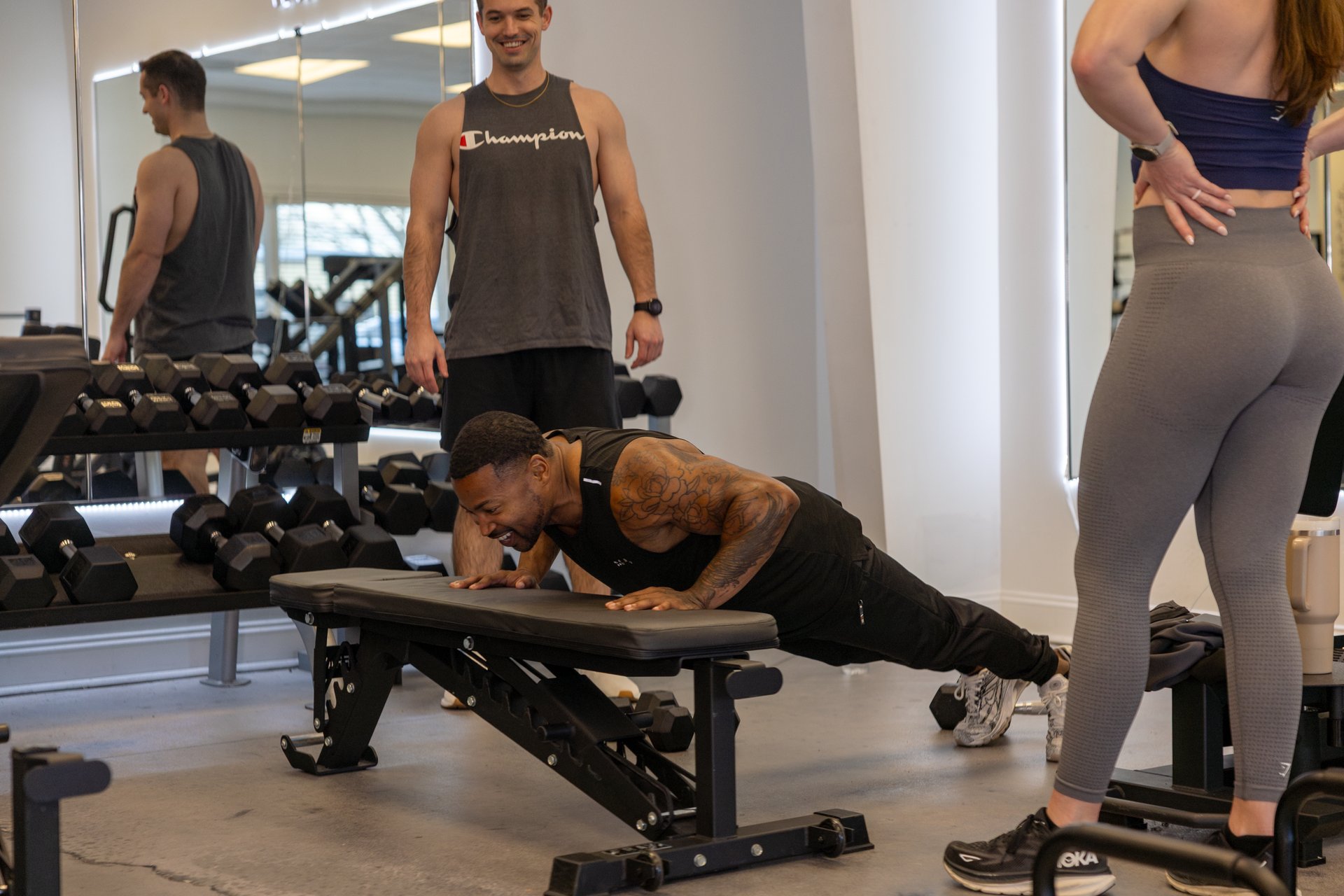 People exercising in a gym; one man does a one-arm push-up on a bench near dumbbells.