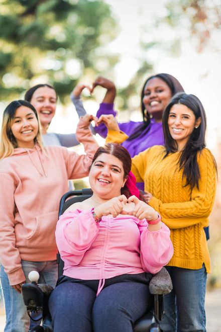 Five friends smiling outdoors, one seated in a wheelchair, wearing colorful sweaters and making a heart shape with their hands