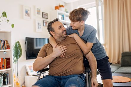 Man in a wheelchair smiling as a child hugs him in a bright living room