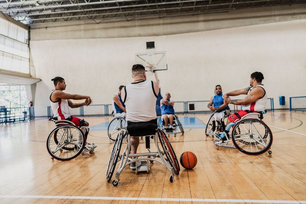 Wheelchair basketball players practice on a gym court with a ball near center.