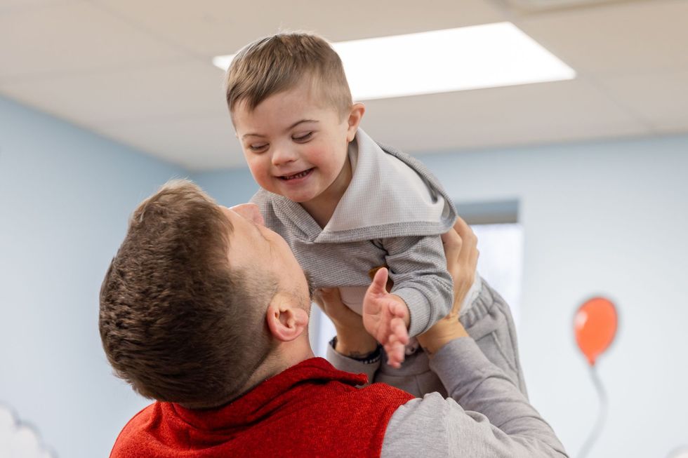 Man holding a smiling child indoors, both laughing and reaching toward each other