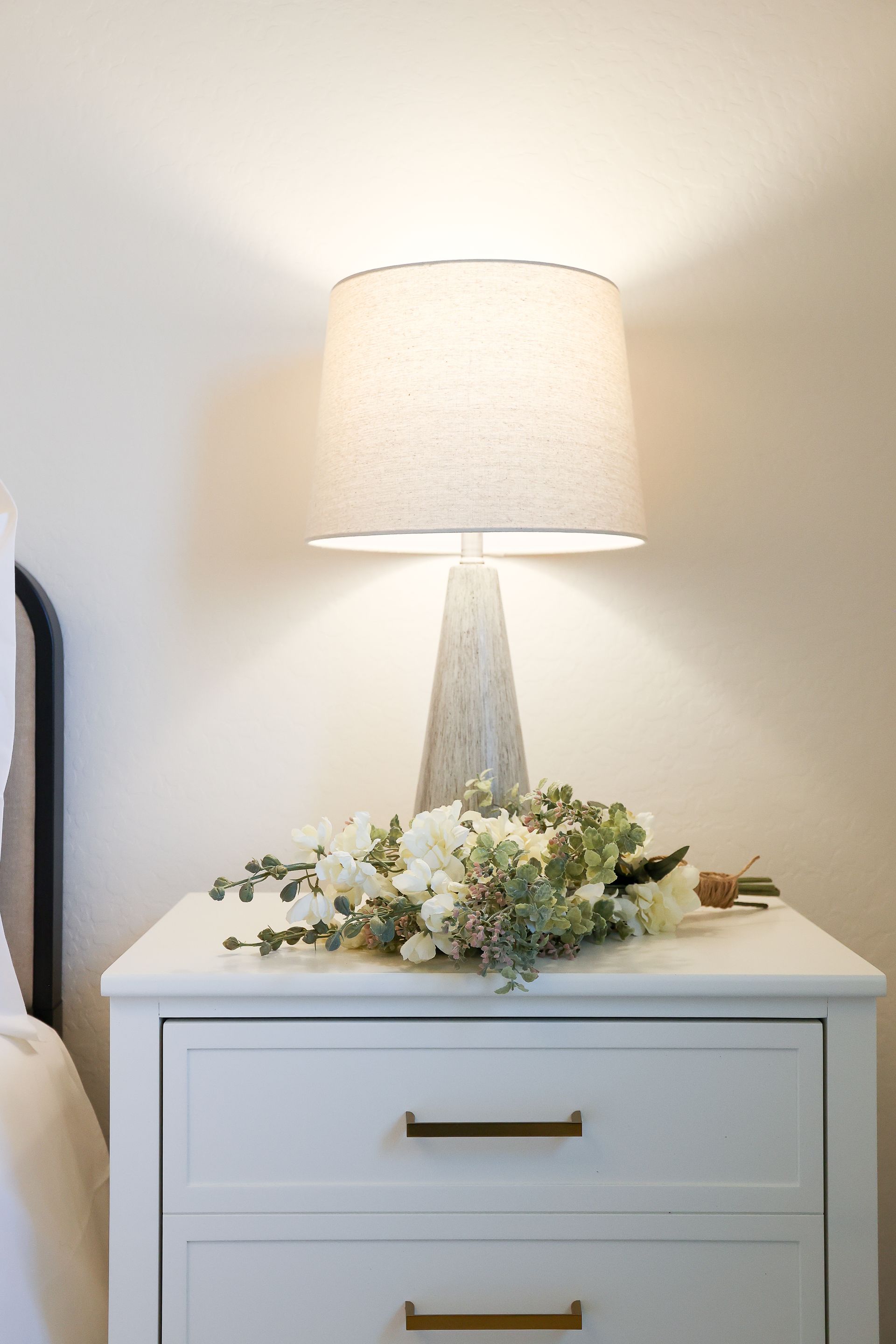 White bedside table with lamp and flowers against a light wall