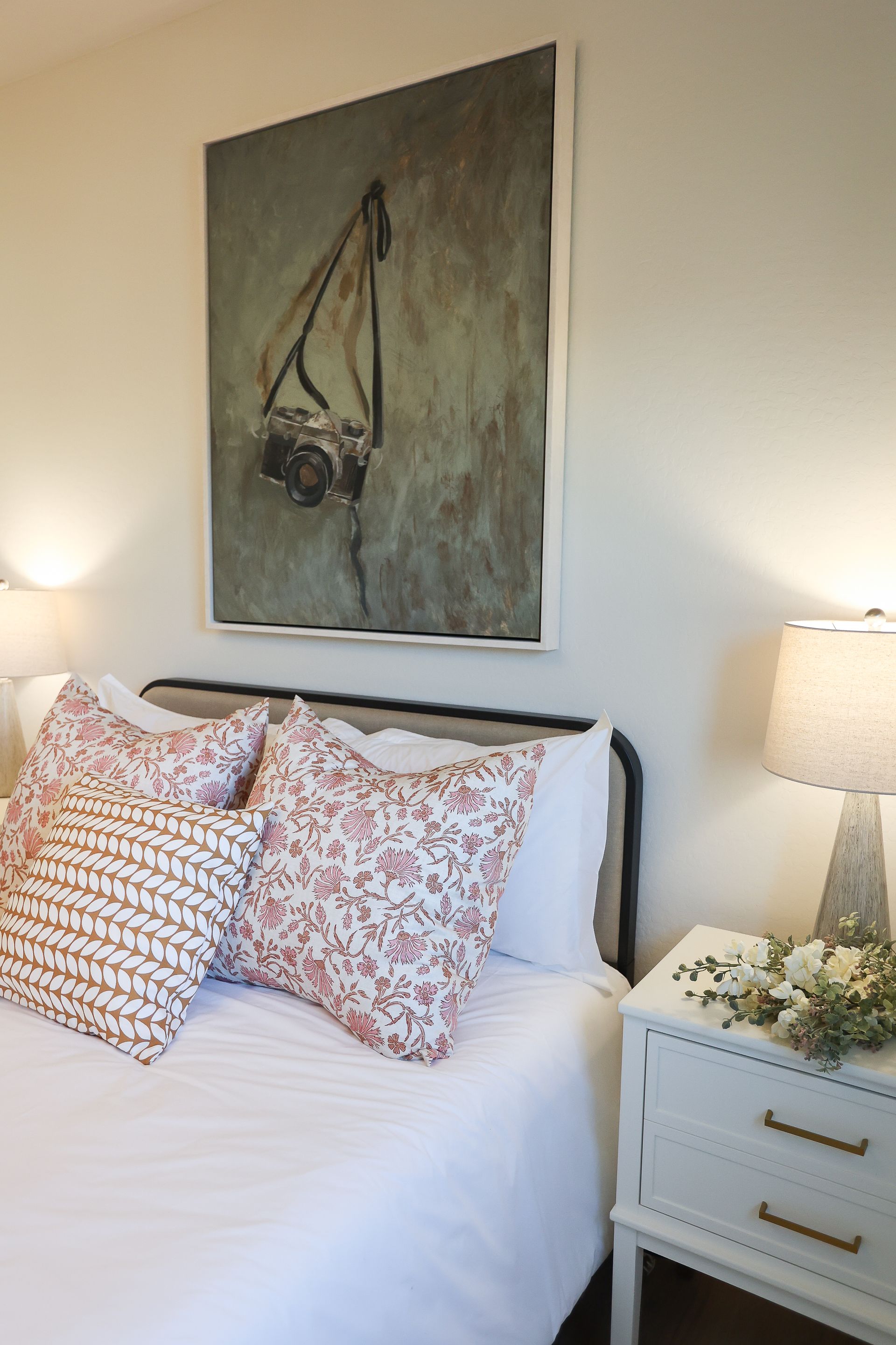 Cozy bedroom with white bedding, patterned pillows, bedside lamps, and a large wall painting above the bed