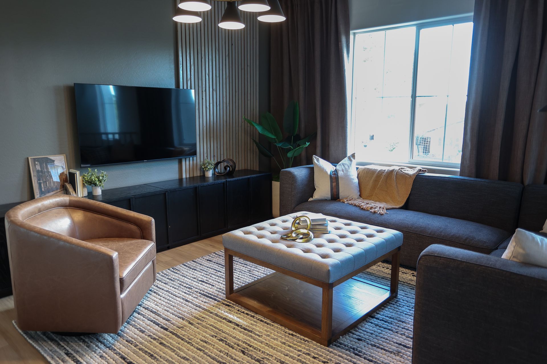 Modern living room with brown sofas, tufted ottoman, TV, and large window with curtains