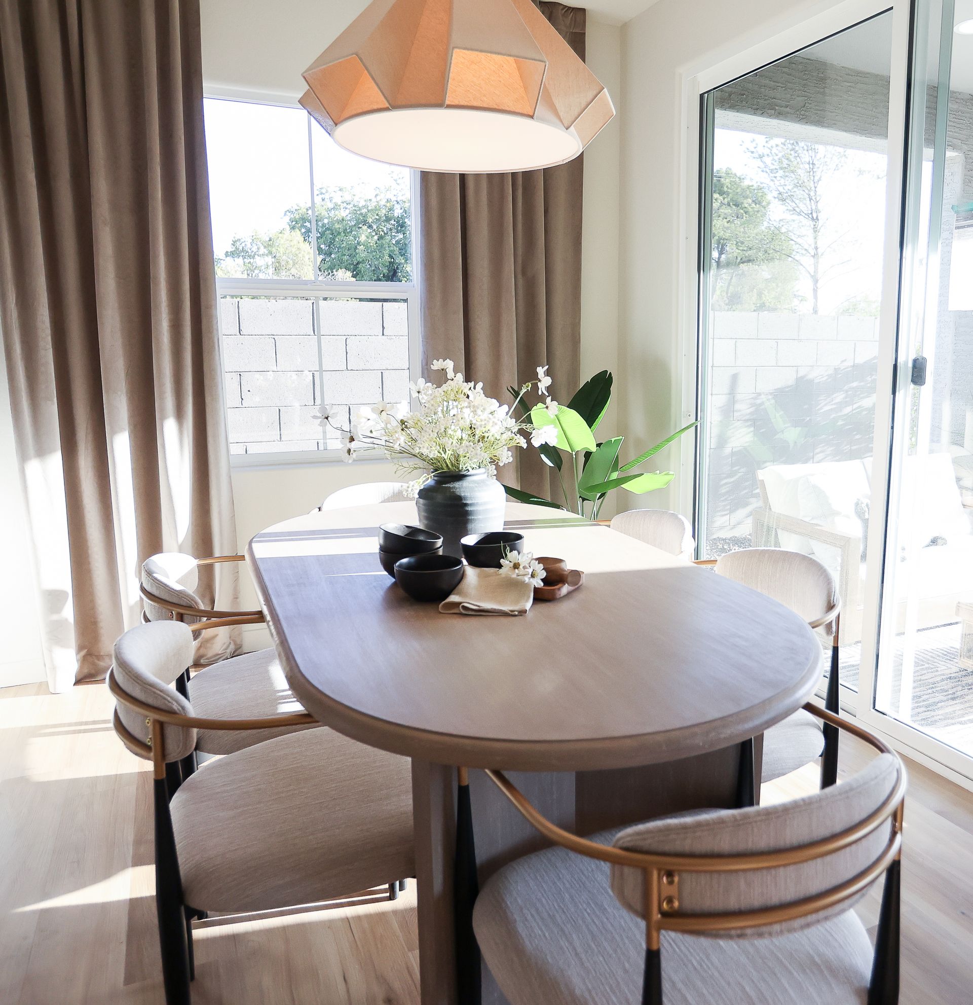 Bright dining nook with round wooden table, cushioned chairs, and a vase of white flowers by large windows.