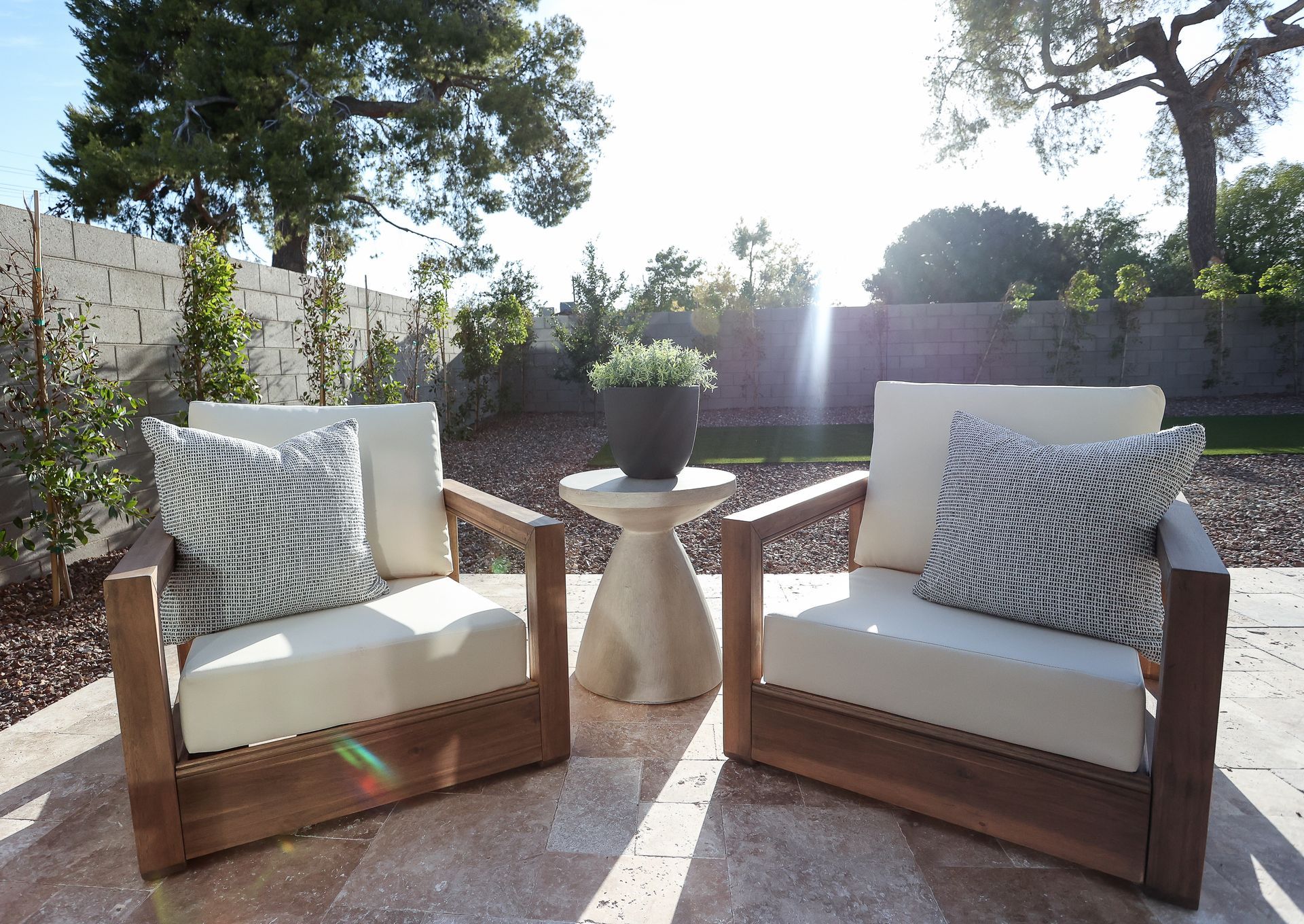Two wooden armchairs with white cushions and patterned pillows on a sunny patio, with a small table between them.
