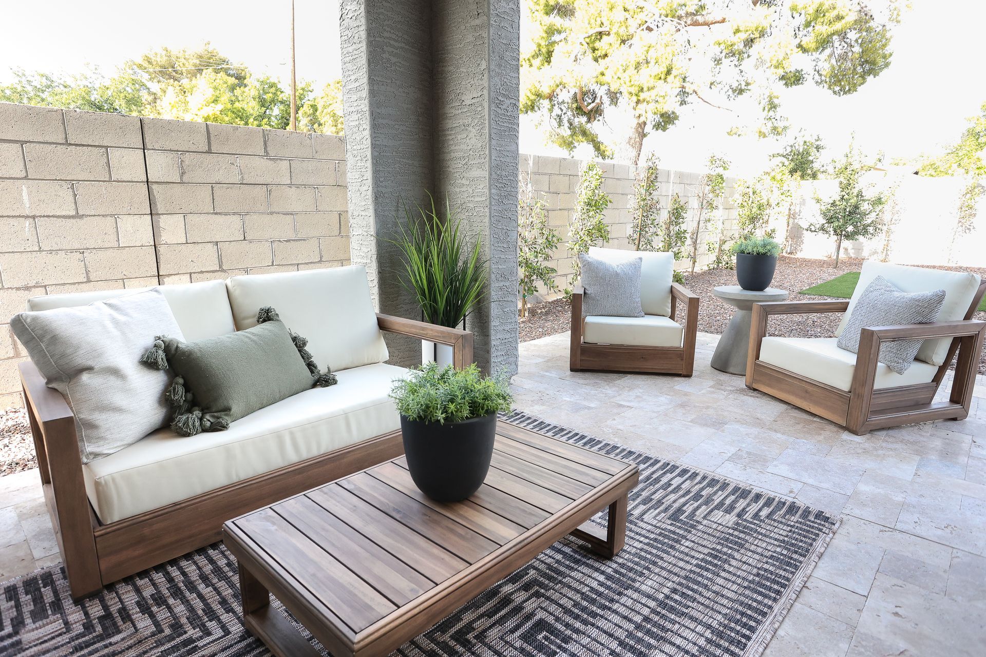 Patio seating area with white and wood sofas, coffee table, rug, and potted plants in bright sunlight