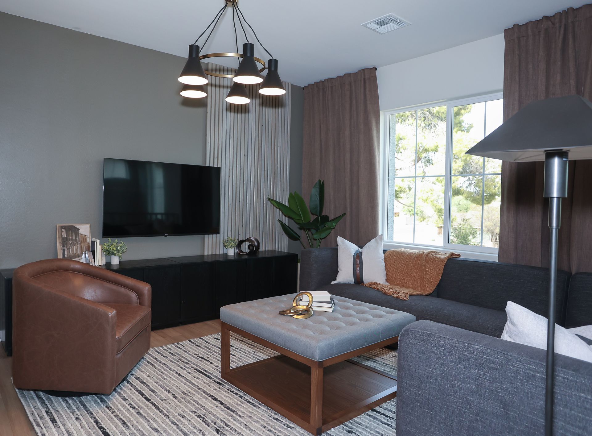 Modern living room with gray sofa, brown chair, TV, coffee table, and large window with curtains
