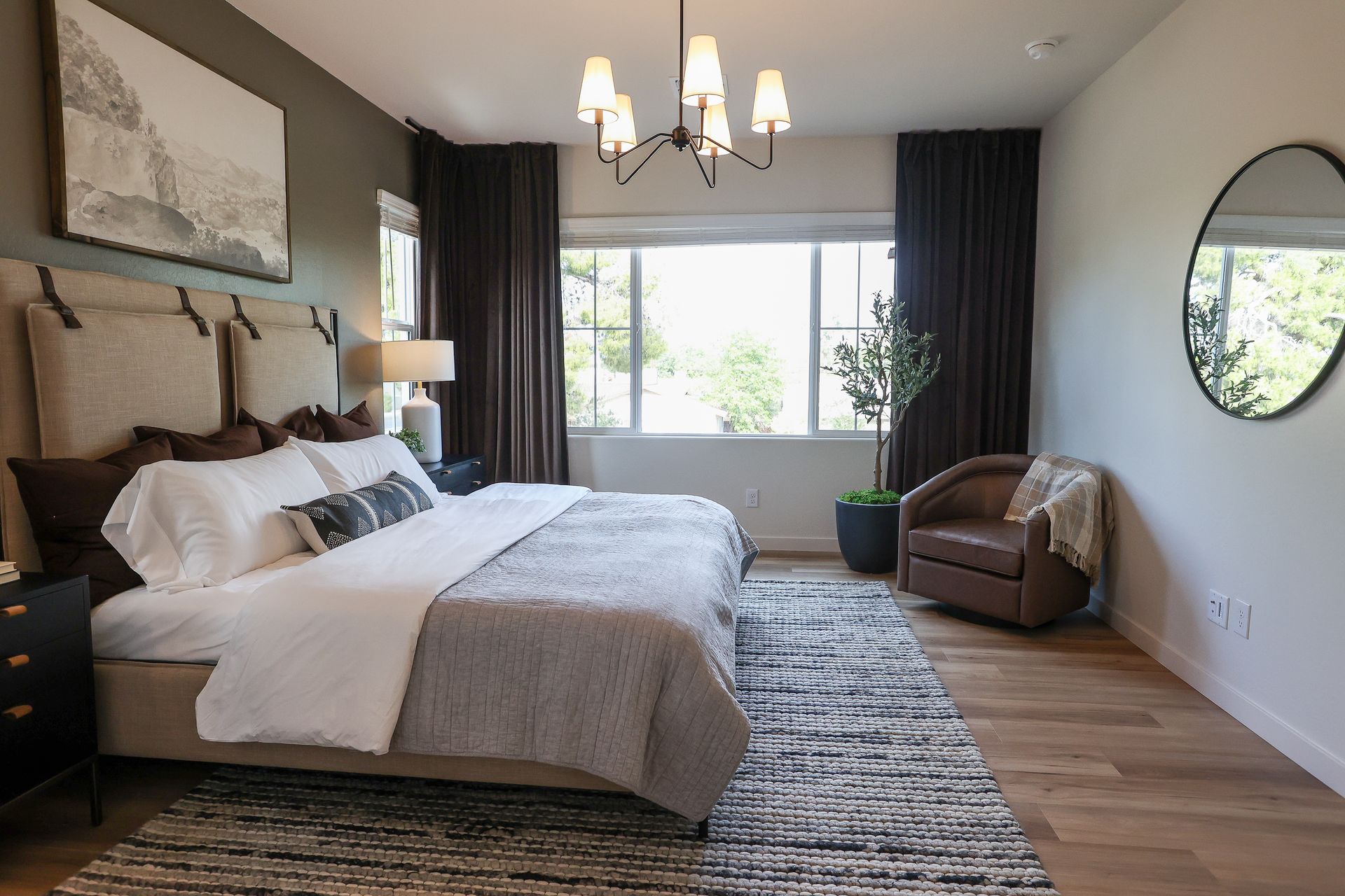 Bright bedroom with king bed, gray chair, chandelier, and large window with dark curtains