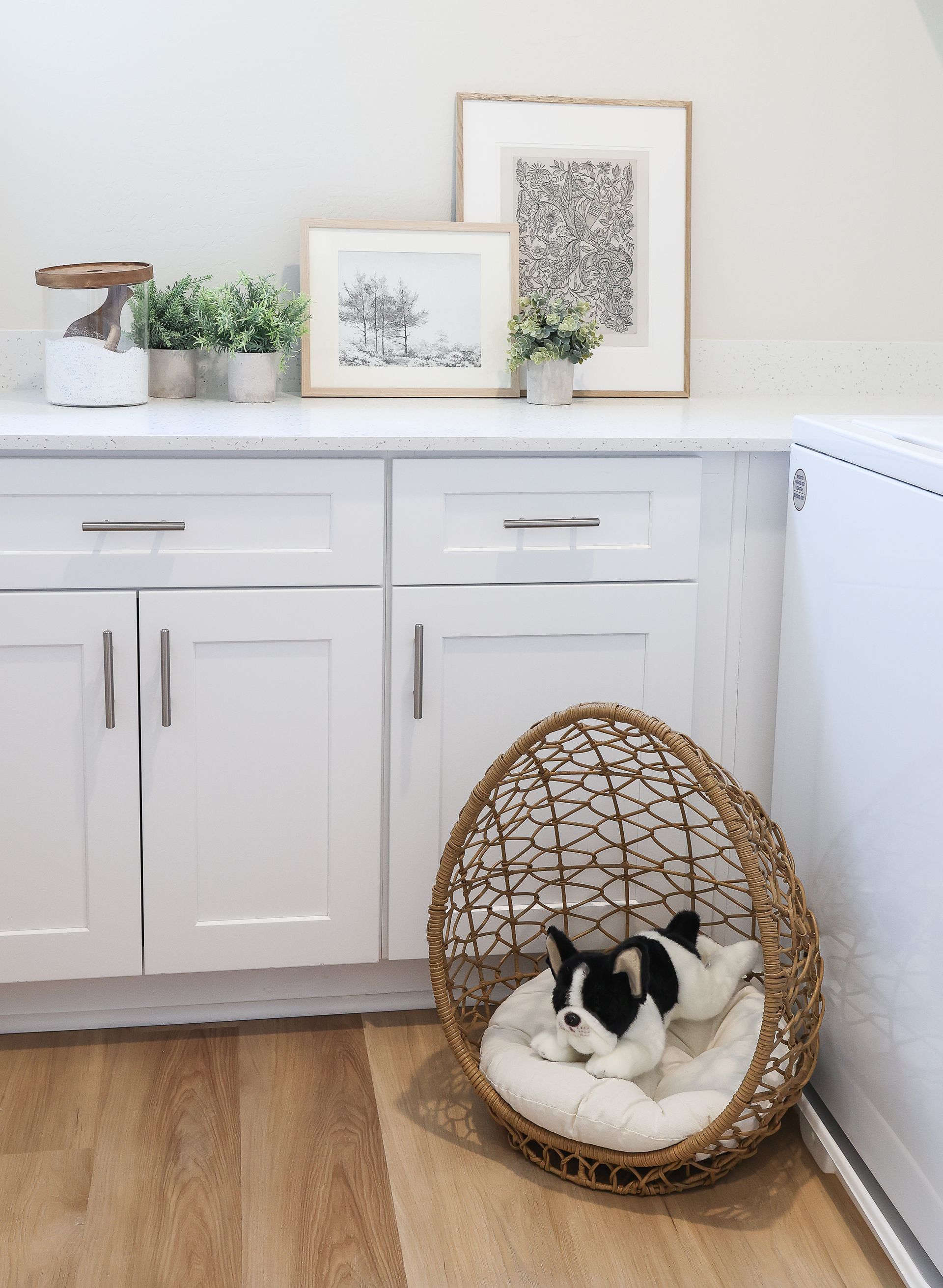 Small black-and-white dog curled in a wicker bed beside white kitchen cabinets on a wood floor.
