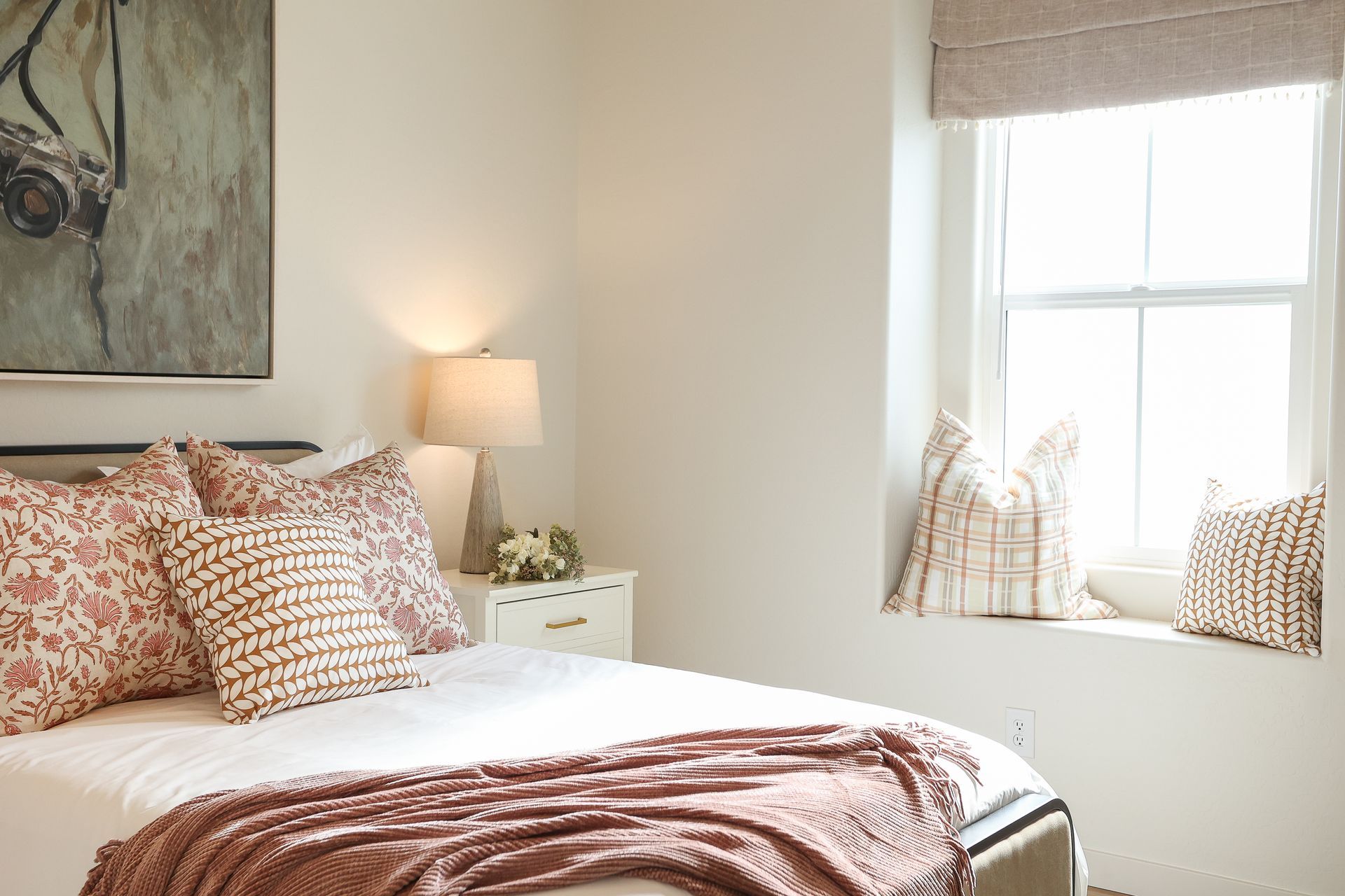 Bright bedroom with bed, floral pillows, lamp, and sunlit window seat with cushions