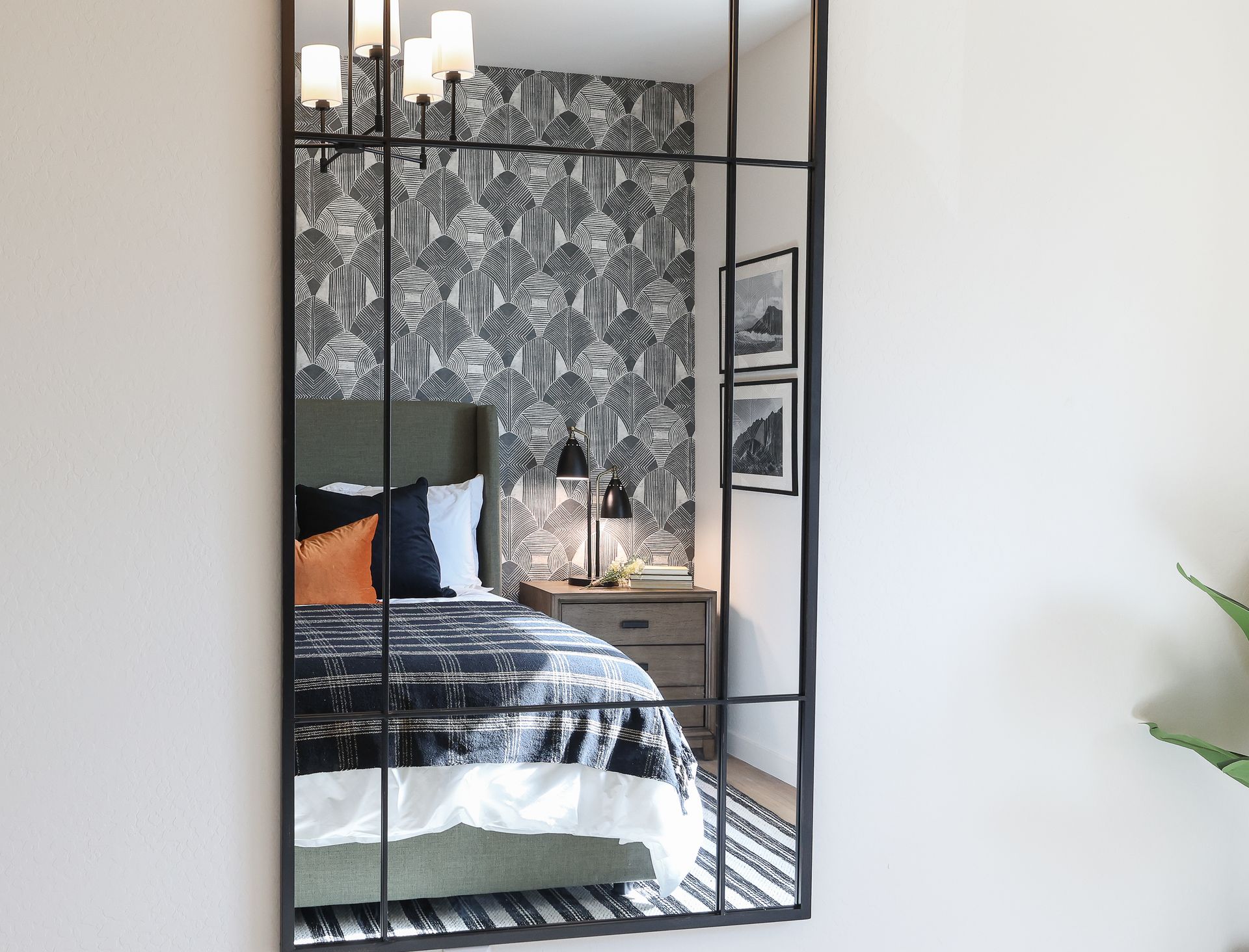 Tall black-framed mirror reflecting a bedroom with a bed, patterned wallpaper, and a bedside lamp