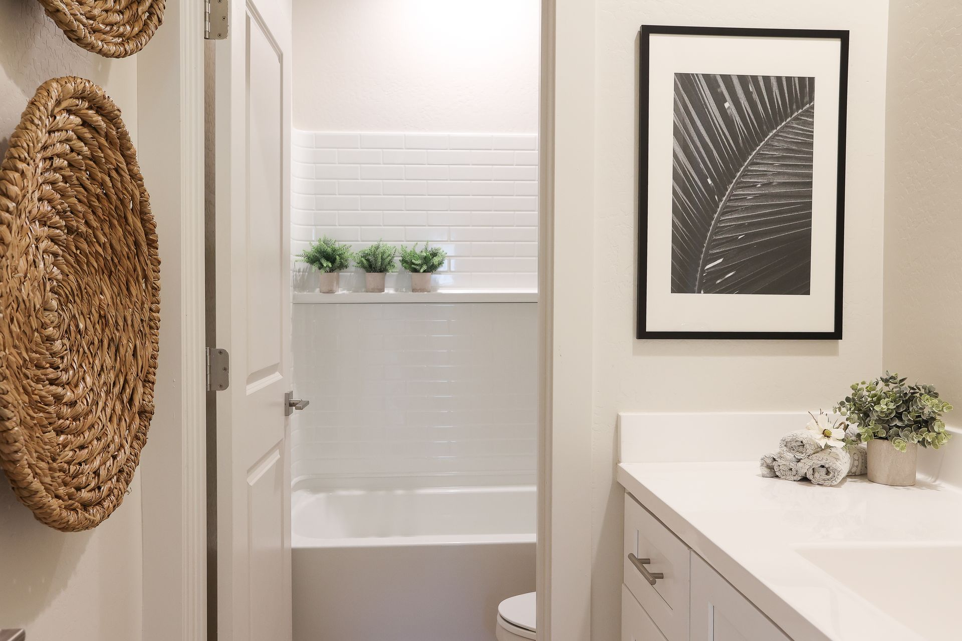 Small bathroom with bathtub, woven wall decor, framed leaf print, and white vanity.