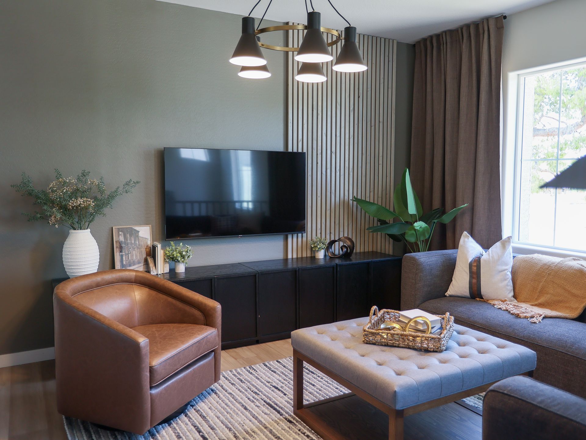 Modern living room with TV, tan chair, gray sofa, and tufted ottoman by a window