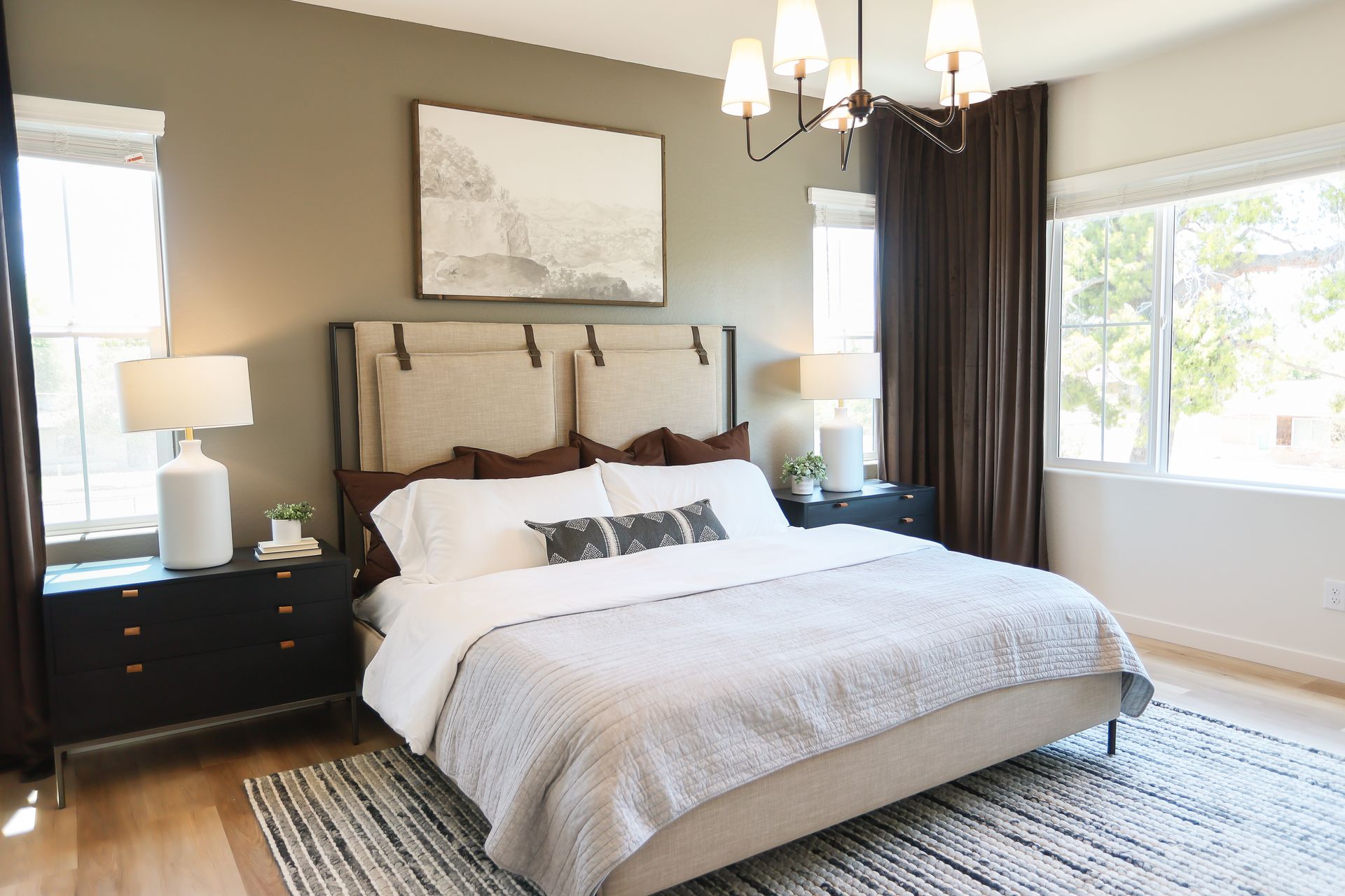 Bright bedroom with a large bed, dark nightstands, a chandelier, and sunlight from two windows.