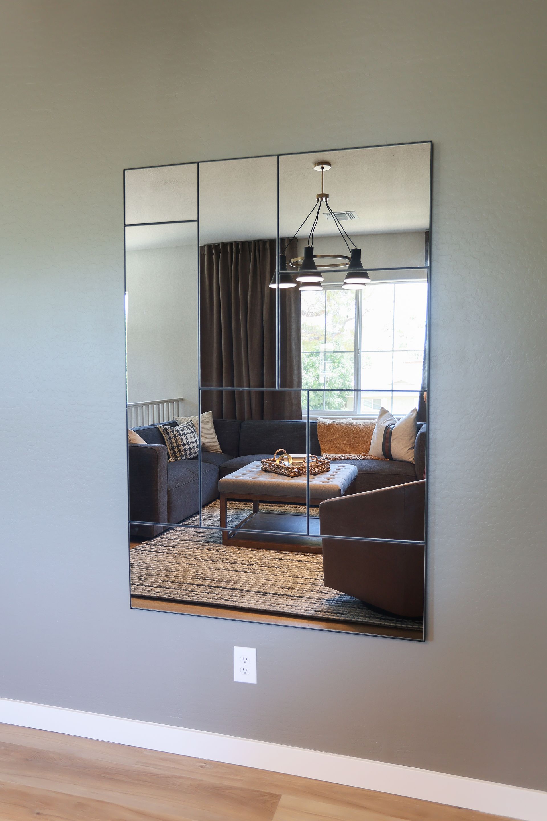 Wall mirror reflecting a living room with a sofa, patterned rug, curtains, and a hanging light fixture.