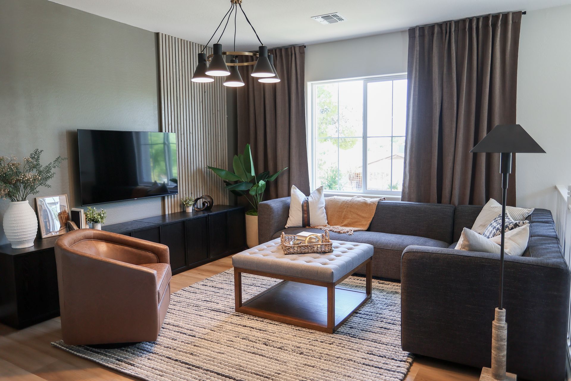 Modern living room with dark sofas, tan chair, coffee table, TV, large window, and pendant light.