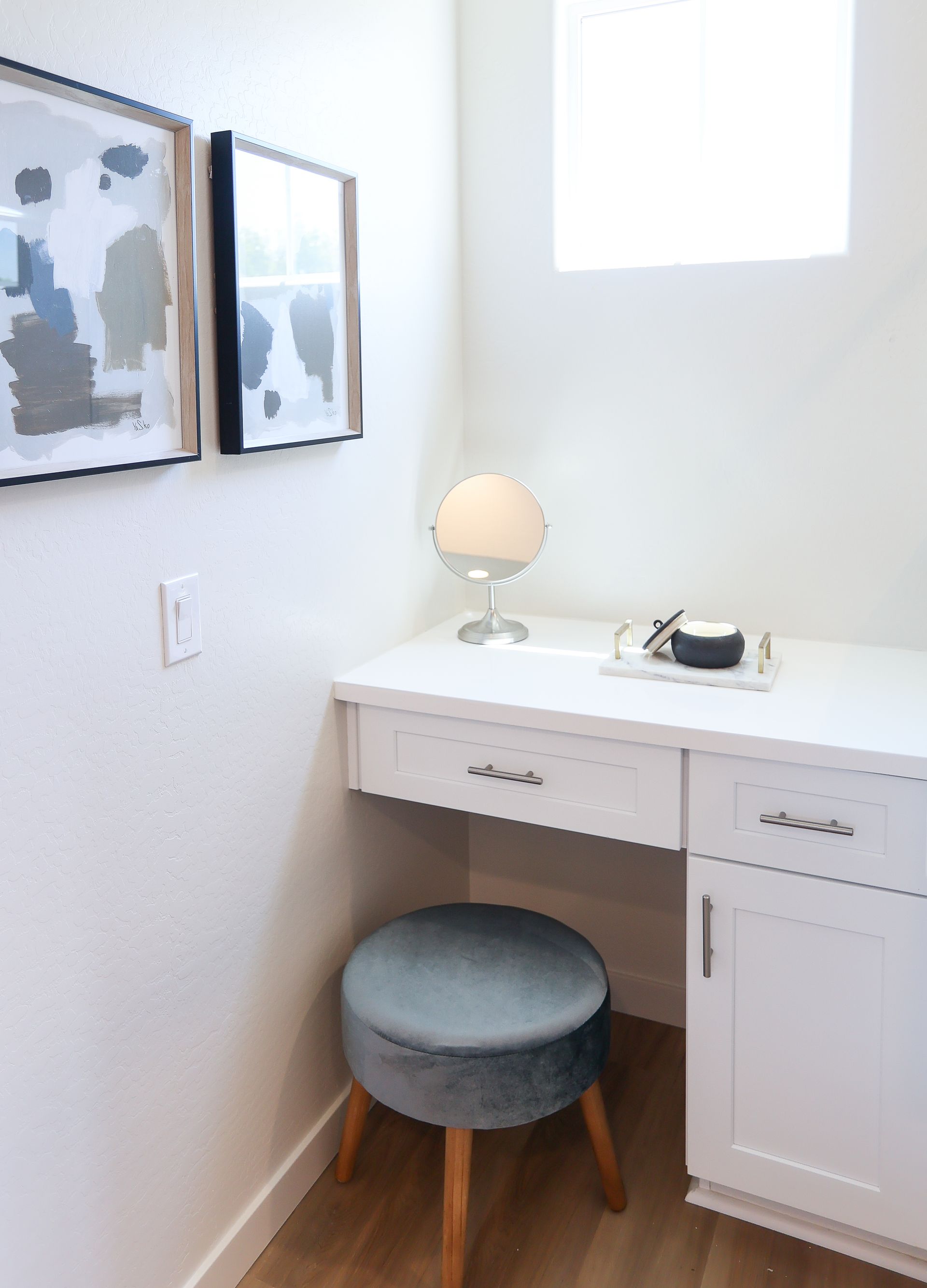Small white vanity desk with a round gray stool, lamp, and phone in a bright corner by framed art.