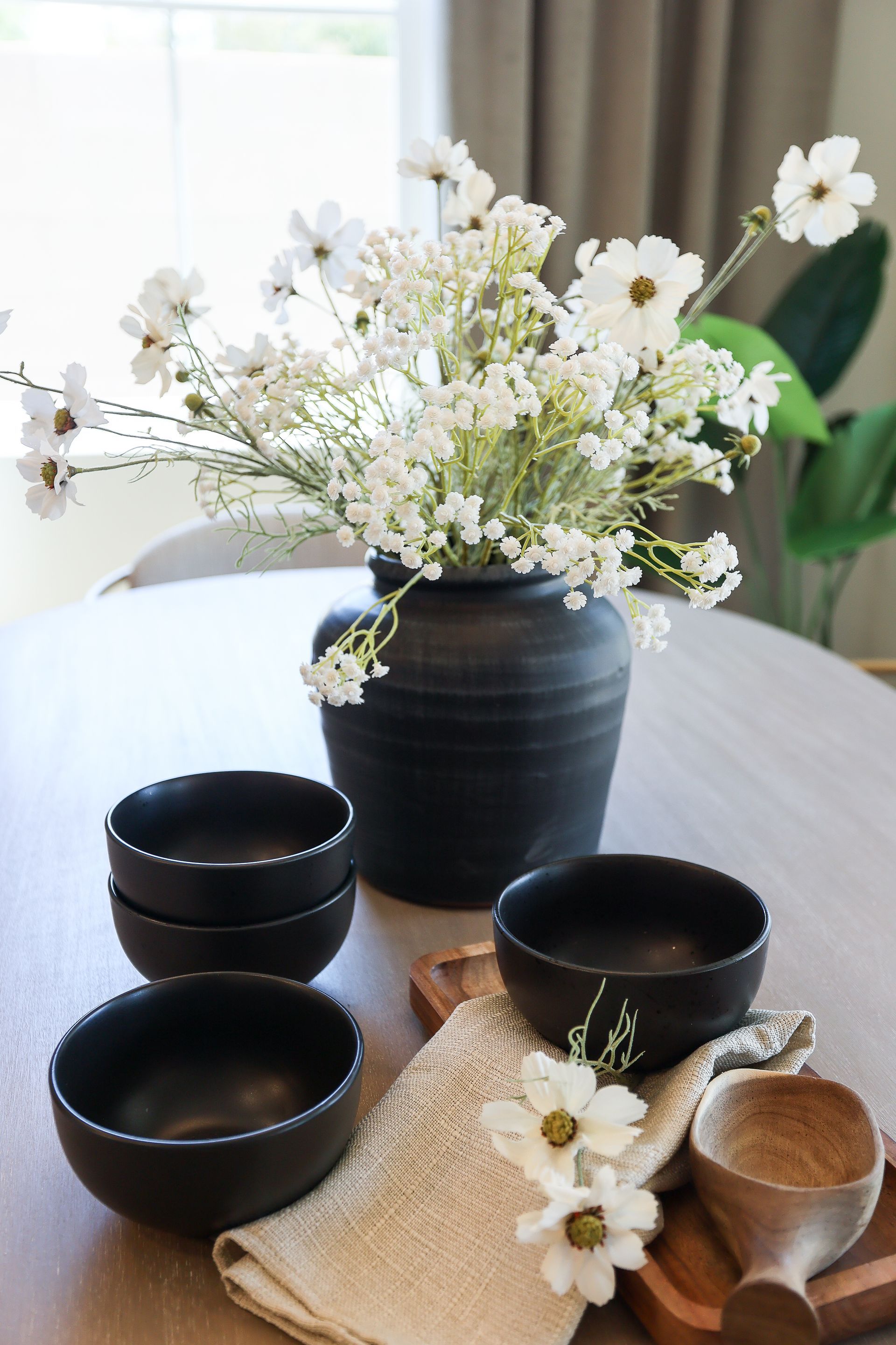 White flowers in a black vase on a table with matching black bowls and a wooden spoon.