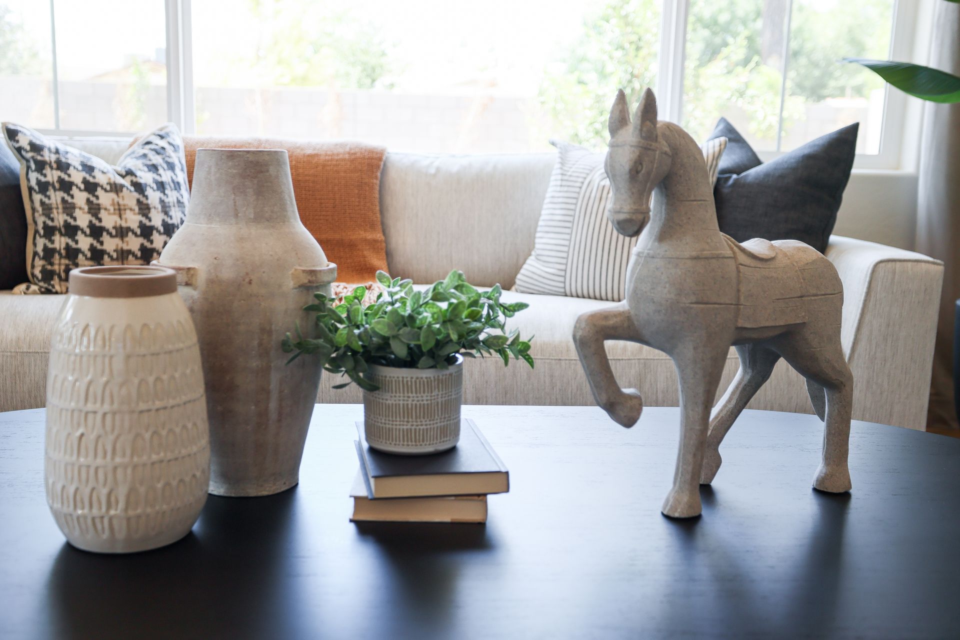 Decorative vases, books, and a small potted plant on a table beside a wooden horse figurine in a bright living room