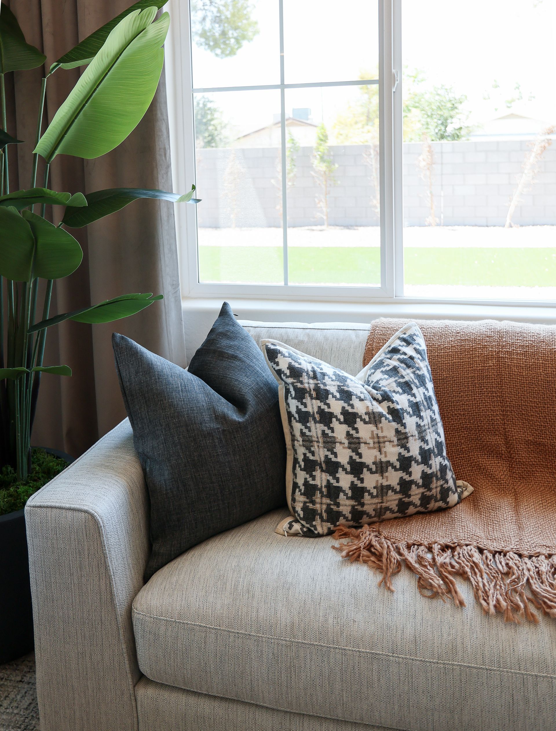 Cozy sofa with dark pillows and a rust throw beside a bright window and potted plant.