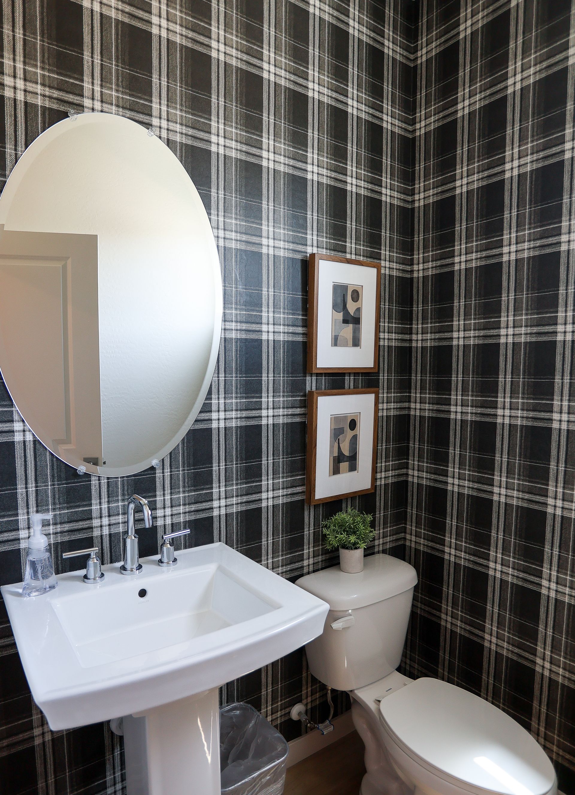 Small bathroom with black plaid wallpaper, pedestal sink, toilet, oval mirror, and two framed pictures.